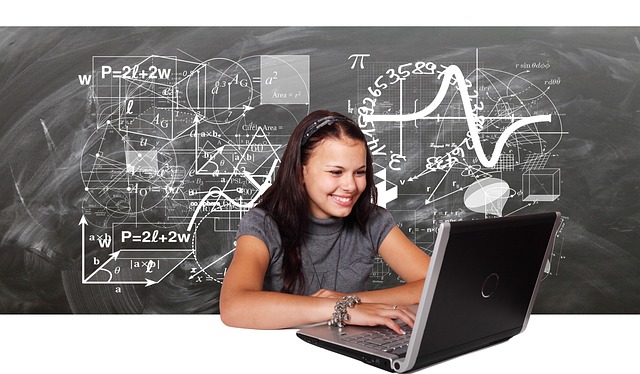 A girl sits in front of a laptop computer in front of a black board filled with mathematical equations